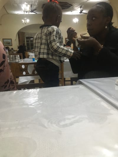 An adult is interacting with a child at a table. The adult, on the right, is dark-skinned with braided hair. The child is on the left and is also dark-skinned. The child is wearing a patterned black and white jacket, with black pants. They are standing at a white table. The interior appears to be a dining or living room, evidenced by chairs, a painting, and light fixtures. The photo is taken indoors, with a slightly rotated perspective.