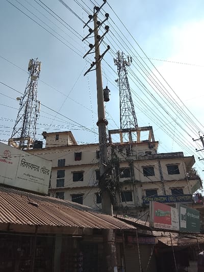 The scene depicts a bustling urban landscape in daylight. An aged, multi-story building is flanked by telecommunication towers. Dense power lines crisscross the sky, connected to utility poles. Commercial signage is visible on the building and nearby shops. A corrugated metal roof of a low structure is in the foreground.
