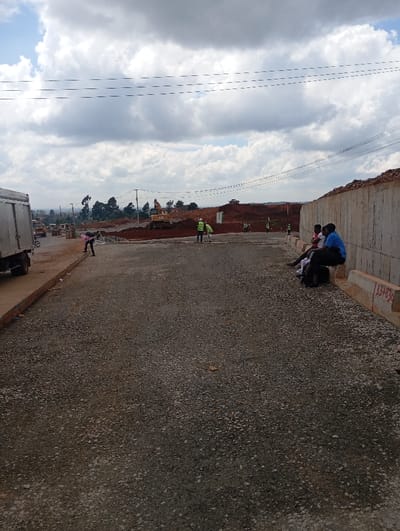 The image displays an active road construction site located in Gitaru ward, Kenya, under an overcast sky. In the foreground, a wide, unpaved road surface composed of grey gravel extends towards the background. To the left, the rear section of a white box truck is partially visible. Near the left edge of the gravel road, a person in dark attire with a pink upper garment is bent over an area of brown earth. In the midground, two individuals wearing yellow high-visibility vests and helmets are standing on the gravel road. Further behind them, a large yellow excavator with an extended boom and bucket is positioned on an elevated section of red excavated earth. Additional construction activity, visible as mounds of red soil, extends into the background. Along the right side of the gravel road, a concrete barrier or retaining wall, marked with red numbers "33+830," runs parallel to the road. Four individuals are seated on this concrete barrier; one wears a blue shirt, and another wears a red shirt. Overhead, multiple utility lines stretch horizontally across the scene. The background reveals a landscape of green trees and disturbed red earth under a cloudy sky.