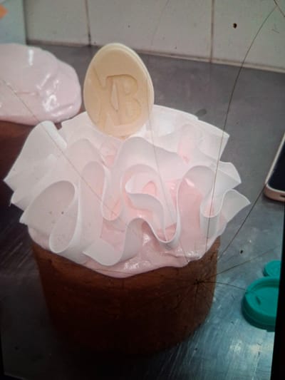 An overhead medium shot captures a cake decorated with pink frosting and white, ruffled paper frills, topped with a light-colored disc embossed with the letters "NB." The cake is positioned on a metallic, reflective surface, possibly a kitchen counter, exhibiting scratches and smudges. To the left of the cake, a partially visible pink blob, possibly meringue or frosting, rests on the surface. In the background, the surface continues, transitioning into a tiled wall. To the right of the cake, two teal lids, likely from containers, are visible. The scene appears to be indoors, possibly in a bakery or kitchen setting in Medvezhyi Ozyora, Russia. No individuals are present in the frame, and the lighting suggests an indoor environment. The image quality is grainy, and several long, dark strands, potentially hairs, are visible across the frame, along with other small dark specks.