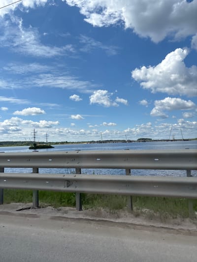 The image displays a wide outdoor landscape scene, likely captured from a road or bridge overlooking a body of water. In the foreground on the right side of the frame, a robust grey metal guardrail runs horizontally. To the immediate right of the guardrail, a section of a light grey paved surface, possibly a road or shoulder, is visible, separated by a strip of green grass and some dirt.
The central and left portions of the mid-ground are dominated by a broad expanse of blue water, which appears to be a river or a large lake, showing subtle ripples on its surface. Across the water, on the distant horizon, a narrow strip of land is discernible, covered with green trees. Several man-made structures dot this distant shoreline, including tall power line towers and what appear to be industrial cranes or other equipment, suggesting infrastructure development.
The upper and left parts of the background feature a clear, bright blue sky filled with numerous white, fluffy cumulus clouds, indicating fair weather. Some thinner, wispy cirrus clouds are also present higher in the sky. The scene is bathed in strong sunlight, suggesting it is daytime, likely mid-day or early afternoon. There are no people visible, and no discernible text appears anywhere in the image. The location context for this scene is Bezborodovo, Russia.