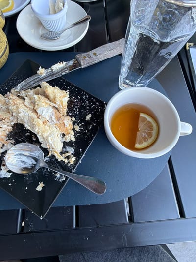 The image shows a table setting, likely outdoors, with various items including a clear glass pitcher, a white mug containing a beverage with a lemon slice, a small dish with a lemon slice, and a black square plate with remnants of a pastry and a blackberry. A knife and spoon, both with traces of cream or icing, are also present. The table is dark, possibly a black plastic or wooden material with a slatted design. The setting appears to be in the city of Glenview, United States of America.