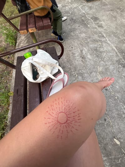 The image shows a close-up of a person's leg with a drawing of a sun on it, located in Garmen, Bulgaria. The person is sitting on a wooden bench. A white purse with a green bottle and keys inside is on the bench next to their leg, along with a pair of pink flip-flops. In the background, a portion of another person wearing green pants and dark sneakers is visible, also seated on the bench. The ground is paved with concrete.