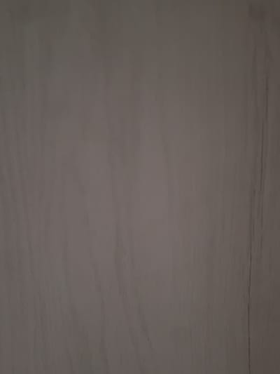 This is a close-up, slightly blurry, overhead shot of a flat, textured surface that appears to be wood or a wood-like material. The color is a muted, neutral tone, somewhere between light brown and gray. There are subtle, darker horizontal lines or grain patterns visible across the surface, especially in the upper portion of the image. The lighting is dim and diffused, with no clear shadows to indicate the time of day. There are no discernible subjects, activities, or emotions depicted. The image is a simple, abstract texture with no identifiable location cues beyond the general appearance of a flat surface, possibly a table or a floor. There is no visible text.