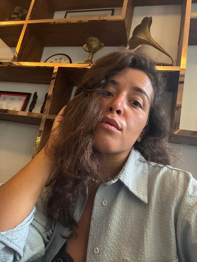 A young woman with long, dark, wavy hair is taking a selfie in what appears to be a room with wooden shelves in the background. She is wearing a light blue collared shirt with textured fabric and pink buttons. One hand is resting on the back of her head, and her left arm is visible. The shelves behind her are filled with various objects, including a framed certificate, small figurines, a trophy, a clock, and what looks like a vintage gramophone horn. The lighting suggests an indoor setting. The image does not provide any direct information to pinpoint the location to Khan Daun Penh, Cambodia.