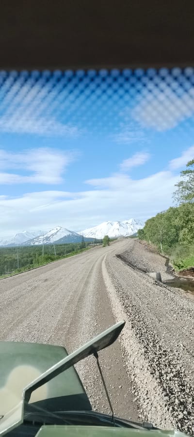 The image captures a view from a vehicle traveling on a gravel road in Termal'nyy, Russia. Snow-capped mountains are visible in the distance, surrounded by green trees and vegetation. The sky is blue with scattered white clouds. The road appears to be under construction or maintenance, with piles of gravel on the side and a culvert visible. The foreground shows a portion of the vehicle's hood and a mirror, suggesting the perspective of the driver. The scene is rural and natural, with evidence of human infrastructure in the form of the road and power lines.