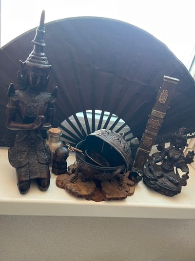 This image displays a collection of dark, intricately carved wooden objects arranged on a white surface, possibly a shelf or countertop, against a backdrop of a large, dark, fan-like object and a window with blinds. The fan appears to be a photography backdrop. The objects include what looks like a statue of a person or deity, possibly in traditional attire, with detailed carvings on its clothing and headdress. Next to it, there's a small glass bottle with a cork stopper. Above these, a carved wooden spherical object, possibly a decorative vessel or mask, is attached by a chain to a wooden base adorned with carved elements. Further up and to the right, a dark, detailed carving depicts what seems to be a mythical creature or figures in a dynamic pose. A long, decorated wooden element, possibly a part of a musical instrument or decorative sword, is also visible.
The lighting suggests it's daytime, with natural light coming from the window. There are no specific weather cues. The overall scene gives a sense of a curated display of artistic or cultural artifacts. The carvings are rich in detail, featuring repeating patterns and figures. Visible text includes "PLAY" on a white object behind some of the carvings, possibly part of a label or packaging. The location context provided is Балаклавский округ, Ukraine.