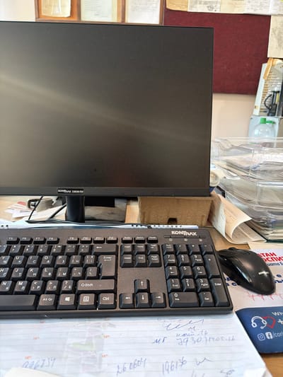 The image shows a close-up view of a desk with a computer monitor, keyboard, and mouse. The monitor has a label that reads "KONTRALX 22B2H/EU". The keyboard is black and appears to be a standard QWERTY layout with Cyrillic characters also visible. A black computer mouse is positioned to the right of the keyboard. In the background, there are stacks of papers and some office supplies. The scene is well-lit, suggesting it is an indoor office or workspace. There are no people visible in the image. The presence of a computer and office supplies indicates a work environment. There are no specific visual cues in the image that would allow for the determination of a specific city, let alone Veliki Preslav, Bulgaria.