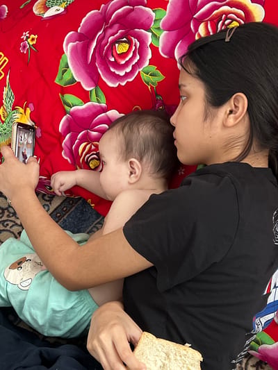 A young woman in a black t-shirt is holding a baby and a smartphone in Kyzylorda, Kazakhstan. The woman is facing left and looking at the phone. The baby is facing away from the camera and looking to the right. The baby is wearing light blue pajamas with a teddy bear print. The woman is holding a piece of bread in her left hand. They are lying on a red blanket with a floral pattern. The floor is covered with a rug.