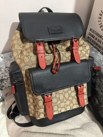 The image shows a Coach backpack resting on a surface. The backpack features a mix of black leather and a tan patterned fabric. It has red leather straps with buckles. The backpack is positioned near a box with text that includes "MEDITERRANEAN CULTURE." No location is specified, and there are no people in the image.