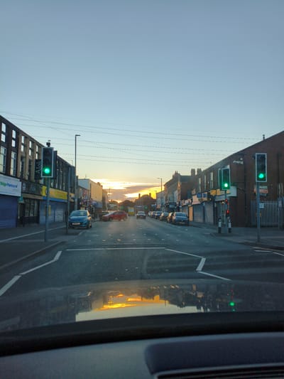 The image shows a view from a vehicle driving down a street at dusk. The street is lined with shops and buildings on both sides, with traffic lights visible. The sky is clear with some clouds on the horizon, and the setting sun casts a warm glow. The road surface is asphalt, with white markings. The reflection of the sky and buildings can be seen on the vehicle's dashboard. This scene is located on Walsall Road in Sandwell, United Kingdom. There are no people visible in the image.