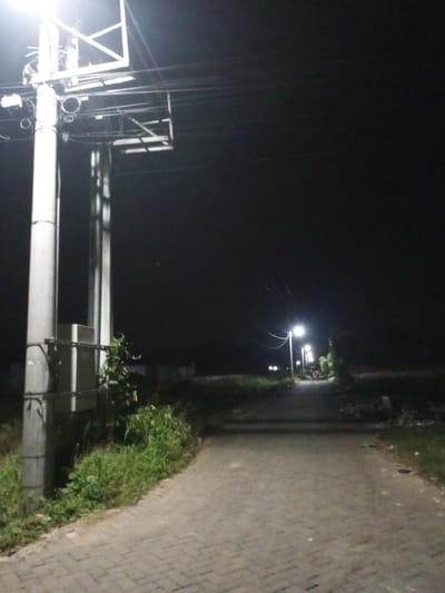 The image depicts a rural road at night in Tibojong, Indonesia. A prominent utility pole with a street light and electrical equipment stands on the left side of the frame, casting a bright light. The road itself is paved with interlocking bricks and curves gently into the distance. Several street lights are visible along the road, illuminating the path ahead.

Green vegetation flanks the sides of the road, suggesting a natural or semi-rural environment. The sky is dark, indicating nighttime. In the distance, a few figures can be seen walking on the road, their forms partially obscured by the low light conditions. The overall atmosphere is quiet and serene, with the artificial lights providing the only significant illumination. There is no visible text in the image. The scene suggests a typical evening in a residential or semi-rural area.