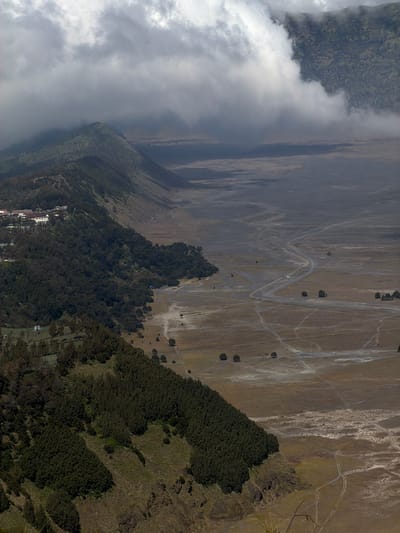 This is an aerial view of the landscape surrounding Cemorolawang, Indonesia. A steep, densely forested slope dominates the left side of the frame, with a small settlement of white buildings visible at the top of the slope. The trees are dark green and appear to be coniferous.
The right side of the image shows a vast, arid plain, likely a caldera floor, characterized by light brown soil and sparse vegetation. Several dirt tracks and a paved road wind through the plain, suggesting human activity. A low fence runs along the edge of the paved road for a significant distance. Isolated trees and small clusters of them are scattered across the plain. In the distance, hazy mountains are visible, partially obscured by thick, low-hanging clouds, which also create shadowed areas on the landscape. The lighting suggests it is daytime, with the sun likely diffused by the cloud cover. There are no visible people in the image.