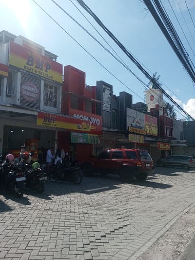 The image displays a daytime street scene in Ban Phonhxavanh, Laos, under a clear blue sky with overhead utility cables. A row of commercial buildings lines the left side, featuring multiple businesses. The leftmost building has a prominent yellow and red sign for "MIE BASO BMP URAT & TETELAN" with an open ground floor. Approximately 7-10 individuals are visible inside or near the entrance of this establishment. Adjacent buildings display signs for "MOMOYO" and "NASI AYAM GEPREK 15.000." In the background, a white tower topped with a windmill-like structure is discernible. The foreground consists of a grey interlocking paver surface used for parking. Five motorcycles are parked near the "MIE BASO BMP" entrance, one with a pink helmet on its seat. A red SUV, possibly a Ford Escape, is parked facing away from the viewer, displaying a partially visible license plate beginning "D 1642." Further down the paved area, a dark sedan is also parked. Shadows are cast by buildings and vehicles, indicating direct sunlight.