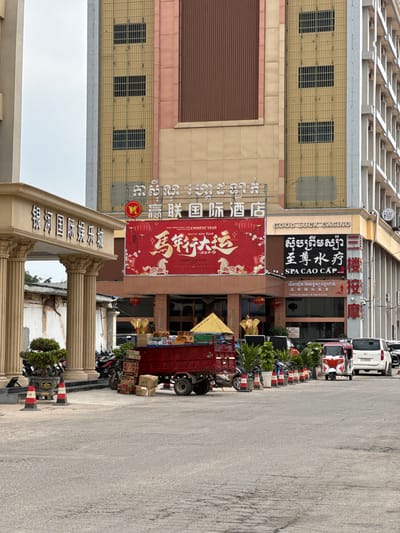 The image depicts a street scene in the Bavet Quarter, Cambodia, showcasing a series of establishments with prominent signage in multiple languages, including Chinese, Khmer, and English. Visible businesses include "Good Luck Casino," "SPA CAO CAP," and a restaurant or hotel named "Ying Lian International." A large red banner advertising the Chinese New Year is also displayed. A red truck is parked on the side of the road, loaded with boxes and goods. Several motorcycles are parked along the sidewalk. The street is paved, and buildings of varying heights and architectural styles line the street. The sky is overcast.