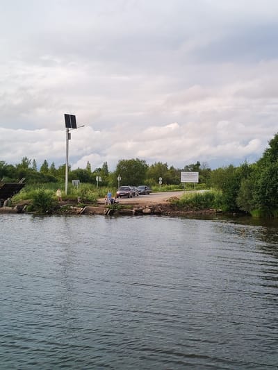 A scenic view of the Kargulino, Russia waterfront features a tranquil body of water in the foreground, reflecting the overcast sky and surrounding greenery. A paved road is visible on the right bank, with several cars parked and driving. A white sign with Russian text stands near the road, partially obscured by lush trees. On the left bank, a person in a yellow dress stands near a solar-powered lamp post with two panels. Several other people are gathered near the water's edge, some appearing to be children. The landscape is dotted with dense green trees and bushes, creating a natural and peaceful atmosphere.