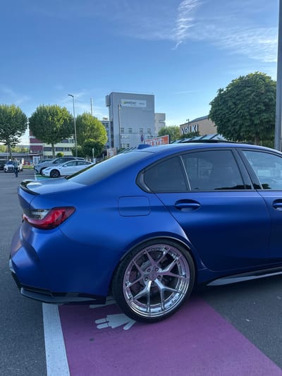The image shows the rear portion of a blue car parked on a purple parking spot marked with a white symbol representing a mother and child. The car has custom chrome wheels and is situated near the street. In the background, buildings are visible, including the "Weber" company building and a building with "Volki" signage. There are trees lining the street. People are seen standing near other cars further down the street. The location is in Volketswil, Switzerland. The sky is blue and clear.