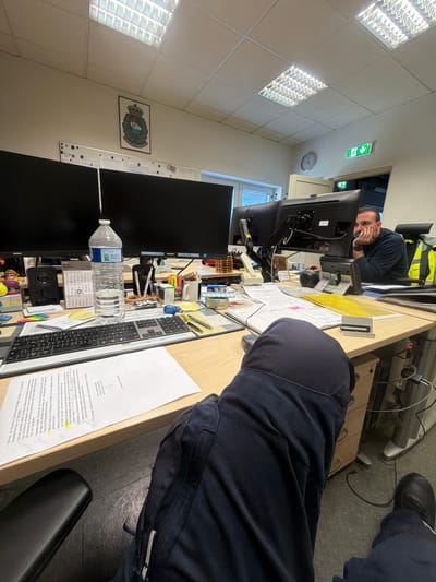 The image depicts an office environment in Grevenmacher, Luxembourg, from a slightly tilted perspective. Two men are present; one is partially visible in the foreground, seated with his back to the viewer, wearing dark blue uniform-like clothing. The other man is seated at an adjacent desk, looking directly towards the camera with a neutral expression, wearing a dark long-sleeved top. He has a beard and appears to be in his 30s or 40s.
The scene is an office filled with typical workspace items. Both men are at desks equipped with computer monitors (including Philips and LG brands), keyboards, mice, and various office supplies like pens, sticky notes, and a stapler. A Vittel water bottle stands on one desk. Papers and documents are scattered across the desks, some with highlighted sections. The background reveals an office ceiling with square fluorescent light fixtures and a green emergency exit sign pointing left. A wall clock shows approximately 1:55. A framed emblem or coat of arms is mounted on a far wall.
The activity appears to be office work, possibly with a brief interruption as one man looks at the camera. The office seems functional and busy.
Visible text on the documents is primarily in German, indicating legal or financial content. Phrases include "Nachweis der monatliche" (proof of monthly), "Zins- und Tilgungsplan" (interest and repayment plan), "Versicherungen" (insurances), "Mitteilung nebst Belegvorlage" (notification along with supporting documents), "Grundsteuer" (property tax), and "Kilometer" (kilometers). The closing of one document reads "Mit freundlichen Grüßen" (sincerely) from "Wischer & Obertig Rechtsanwälte" (Wischer & Obertig Lawyers), suggesting the setting is a law firm or a department dealing with legal matters. The water bottle clearly shows the "Vittel" brand.
