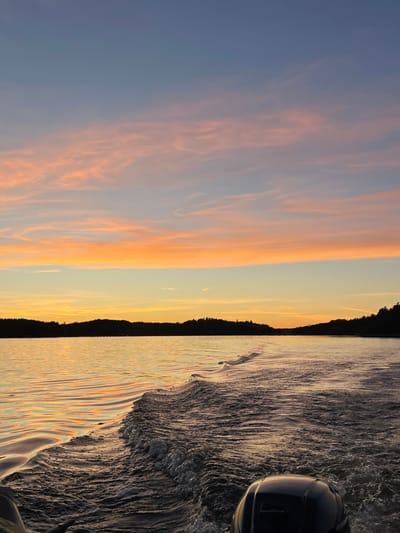 The image depicts a view from the stern of a moving boat on a body of water within Ekerö kommun, Sweden, during either sunset or sunrise. The sky occupies the upper two-thirds of the frame, displaying a gradient from deep blue at the top, transitioning through lighter blue, to horizontal bands of vibrant orange and pink altocumulus clouds above the distant horizon. The horizon features a dark, undulating silhouette of land, indicative of a forested shoreline. The water in the foreground and mid-ground reflects the warm orange and golden hues of the sky, creating shimmering patterns. A dark blue or black outboard motor is partially visible in the bottom-right corner. A prominent, turbulent white wake trails from the boat's stern, extending diagonally from the bottom-center towards the middle of the frame, indicating the boat's forward motion. No individuals are visible.