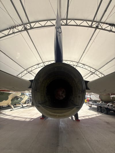 The image shows the rear of an aircraft engine under a white-netted, arched structure. The engine exhaust nozzle is prominently displayed, revealing the interior details. Part of the aircraft's tail fin and fuselage are visible. In the background, other military vehicles and equipment are present, suggesting a museum or storage facility. The setting appears to be indoors, with bright lighting illuminating the engine and the surrounding area. The image is rotated and the camera is positioned behind the engine.