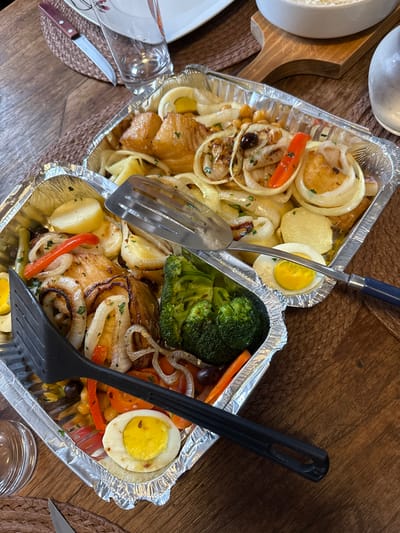 The image displays two aluminum foil takeout containers filled with a prepared meal, presented on a wooden table. The primary subject is a Portuguese-style dish, likely Bacalhau à Brás or similar, containing large pieces of roasted cod fish, sliced onions, roasted potato slices, red bell pepper strips, whole chickpeas, and black olives. One container also includes florets of steamed or roasted broccoli. Several halved hard-boiled eggs are distributed among the food. A silver metal serving spatula with a blue handle rests on the upper container, while a black plastic serving spatula lies across the lower container. In the upper left background, a portion of a white plate, a clear drinking glass, and a knife with a brown wooden handle are visible on a brown woven placemat. A white ceramic bowl is partially visible in the upper right. The scene is set in Porto Alegre, Brazil.
