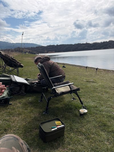 The image displays an outdoor fishing scene at a lakeside location near Cherna Gora, Bulgaria, under a partly cloudy sky. A male individual is seated in a dark green, adjustable fishing chair, leaning forward and engaging with items inside a large, open, dark green tackle bag positioned on the grass in front of him. He is wearing a camouflage-patterned hooded jacket and dark trousers. To his left, another large, dark green fishing chair or bedchair is partially visible. The bare forearm and hand of a second individual, holding a red object, are visible in the far left foreground.
On the grass in the immediate foreground, a small, rectangular dark green tackle box contains a silver knife, a blue tool, and a yellow container with small fishing tackle items. A silver mug is attached to the lower leg of the main subject's fishing chair. Along the water's edge to the right, at least three fishing rods are deployed from rod pods or banksticks. The body of water, likely a lake or reservoir, is calm with grey-blue tones, reflecting the sky. The far bank consists of a treed shoreline backed by rolling hills. Several indistinct figures are visible further along the distant bank, indicating other individuals engaged in fishing. The immediate ground is covered in short green grass.