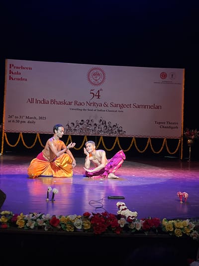 The image shows a performance at the Tagore Theatre in Chandigarh, India, located at the coordinates 30.733, 76.789. Two male dancers are performing on the stage. The dancer on the left wears an orange and gold costume and is in mid-gesture, while the dancer on the right wears a purple and gold costume and is sitting with his right hand on his chin. Both dancers are bare-chested and have dark hair. The stage is decorated with garlands of flowers and a black cable and cell phone lay in front of the dancers. A banner behind the dancers advertises "All India Bhaskar Rao Nritya & Sangeet Sammelan," the "54th Unveiling the Soul of Indian Classical Arts,"  and "26th to 31st March, 2025 at 6:30 pm daily." The banner also lists the venue as "Tagore Theatre Chandigarh." The backdrop behind the banner is a dark, blurred curtain. The lighting is dim, casting a bluish hue over the stage.