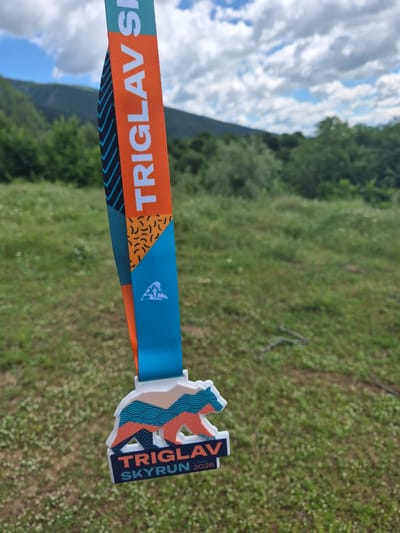 A close-up, shallow depth-of-field shot shows a medal for the "Triglav Skyrun 2026" event hanging from its ribbon. The ribbon is orange and turquoise with a black chevron pattern and abstract orange and black shapes. The medal itself is shaped like a bear with colorful mountain-like patterns and the event name and year prominently displayed. The background is a blur of green grass with scattered white flowers, and distant green trees and hills under a cloudy blue sky. The location is identified as the city of Tazha, Bulgaria. There are no people or animals visible in the image.