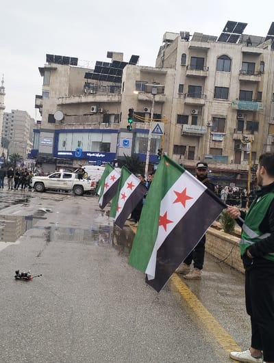 A street scene shows individuals holding Syrian flags. A drone sits on the wet pavement near the flag bearers. A white vehicle with writing on its side is parked nearby, surrounded by other people. Buildings line the background, many adorned with solar panels. A tall minaret is visible in the distance, adding to the urban setting.