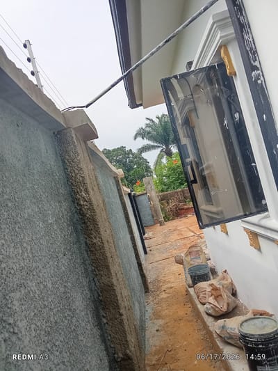 The image depicts an outdoor construction scene in Nawfia, Nigeria. The foreground is dominated by a rough, textured concrete wall. A fence with electric wiring runs along the top of the wall. To the right, a white building with decorative trim surrounds an open window with dark tinted glass. Construction materials, including bags of cement and buckets, are scattered on the ground and a ledge in front of the building. In the background, lush green trees and vegetation are visible, suggesting a tropical environment. A palm tree stands out. The ground appears to be made of dirt and gravel, indicating ongoing work. The sky is overcast, suggesting a cloudy or possibly rainy day. The time stamp on the image reads "06/17/2026 14:59". The text "REDMI A3" is visible in the bottom left corner. No people are visible in the image, but the presence of construction materials suggests human activity is taking place or has recently occurred.