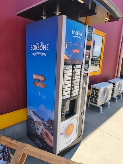 This is a full, eye-level, outdoor shot of a coffee vending machine in the city of Unknown, Bulgaria. The machine is tall and white, with a large blue graphic on its side. The graphic features the text "CAFFÈ BORBONE" and an image of a snowy mountain landscape with a hand holding a cup of coffee. To the right of the vending machine, two white air conditioning units are visible. The background is a red wall and a yellow awning. The ground is paved with concrete. A wooden pallet is in the foreground, with some bags and debris on it. A grey van is visible through a yellow-framed window in the red wall.