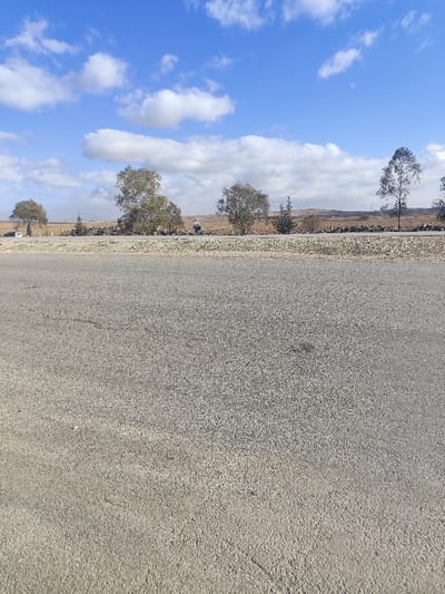A road dominates the foreground, its surface textured with fine gravel. Beyond the road, a low barrier separates it from a landscape dotted with sparse trees and shrubs. In the distance, arid fields stretch towards the horizon. A solitary figure stands near the center, possibly a pedestrian. The sky above is bright blue with scattered white clouds. The overall scene suggests a rural setting with open spaces and a sense of stillness.