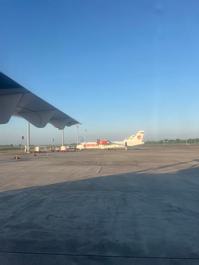 The image shows a Wings Air ATR 72-600 aircraft parked on an airport apron. The aircraft is viewed from the window of another aircraft, with a partial view of its wing in the lower left corner of the frame. Ground support equipment, including service vehicles and cones, surround the Wings Air aircraft. The apron is made of concrete with visible expansion joints. In the background are trees and what appears to be the urban environment of the city of Baji Mangngai, Indonesia, under a clear, blue sky. The lighting suggests it is daytime.