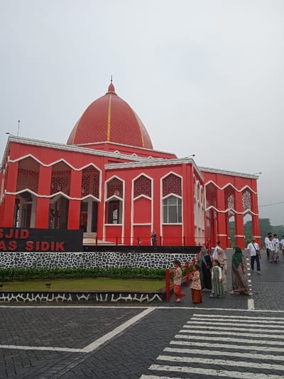 An external view depicts the Masjid As Sidik, located in Tanjungarum, Indonesia, under an overcast sky. The prominent structure is a large mosque, predominantly colored red with white decorative trim and geometric patterned window screens. It features a grand, red tiled dome with golden accents, topped by a pointed finial. A black sign with white lettering displaying "MASJID AS SIDIK" is positioned in front of the mosque, atop a low, patterned stone wall with black and white tessellations, bordering a strip of green grass and small shrubs. In the foreground, a dark gray paved area, possibly a road or plaza, exhibits white painted crosswalk stripes and directional lines. Multiple individuals are present on this paved area and near the mosque. In the right foreground, a group of approximately eight people, including men in white shirts and sarongs, and women wearing hijabs and various modest attire, are walking towards the right side of the frame. In the left-center foreground, two young girls in patterned brown and orange outfits walk near the grassy area. A few additional figures are visible near the elevated entrance of the mosque in the mid-ground. Green foliage is visible in the background on the far right.