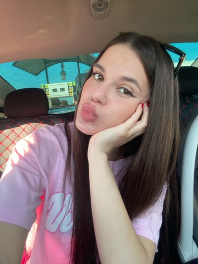 A young woman with long, dark brown hair is taking a selfie inside a car. She is wearing a pink t-shirt with a white logo. Her left hand is resting on her cheek, and her lips are pursed. She is wearing a nose ring and has eyeliner on her eyes. The interior of the car is visible, including a headrest with red stitching and a window reflecting a building and sky. The image does not contain any information that can determine the location in the city of Herasymivka, Ukraine.