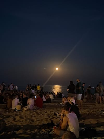 The image depicts a nighttime scene on a beach in Canet de Mar, Spain, with a full moon illuminating the sky and reflecting on the water. A crowd of people is gathered on the sand, some sitting and others standing. The atmosphere appears to be social, with individuals engaged in conversation and enjoying the evening. The sandy beach stretches towards the sea, and the water is calm. The sky is a deep blue, characteristic of nighttime, with the moon as the primary light source. The moon is positioned in the upper-middle section of the image, casting a bright glow. The people in the foreground are closer to the camera, with some individuals clearly visible. They are dressed in casual attire, suitable for a beach setting. The middle ground shows more people spread across the beach, and in the background, more figures are visible along the shoreline. The overall impression is that of a relaxed gathering on a warm evening, with the natural beauty of the moonlit beach as the backdrop.