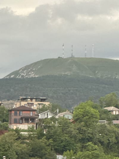 The image depicts a residential area nestled against a steep, tree-covered mountainside. The perspective is from a slightly elevated vantage point, looking across buildings and trees towards the natural landscape. Several buildings are visible, including a prominent brick structure with multiple windows and balconies, suggesting multi-story dwellings or apartments. Other buildings, possibly houses with pitched roofs, are interspersed with dense green foliage, indicating a well-established neighborhood. In the distance, on the left side of the frame, faint vertical structures resembling communication towers or possibly industrial stacks rise from a hazy, overcast sky. The sky itself appears heavy with clouds, suggesting a dull or overcast day. The overall scene conveys a sense of a community situated within a natural, mountainous environment. There is no visible text in the image.