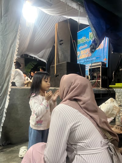 In Wedarijaksa, Indonesia, a young girl with dark hair stands clapping her hands in front of an adult woman wearing a light brown hijab and a striped dress. The girl is wearing a light-colored shirt with a floral pattern and blue pants. Behind them, a large speaker system is visible, along with a blue banner featuring Arabic script and text that appears to be an announcement. The scene is set outdoors under a temporary tent or canopy, with white fabric draped overhead. The ground appears to be concrete. Several other people, some wearing peci hats, are visible in the background, possibly participating in or observing an event. A white clog is lying on the ground near the girl's feet.