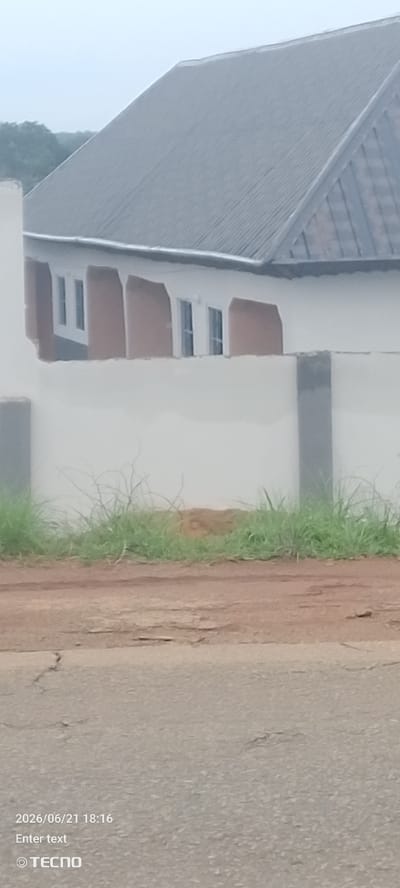 The image depicts an exterior view of a partially constructed building in Gumel, Nigeria. The building features a dark gray corrugated metal roof, white walls, and several rectangular windows. A light-colored wall encloses the property, with a section of unpaved ground and grass visible in front of it. The foreground shows a cracked asphalt road. The timestamp in the lower left corner reads "2026/06/21 18:16" and a TECNO logo is present.