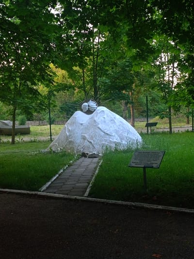 In the city of Urozhaynyi, Russia, a monument is situated in a grassy park area. The monument is a large, white, sculpted rock formation with two human faces emerging from the top, depicted in profile and touching. Above the faces is a circular element with rays, resembling a sun. A paved walkway leads up to the monument. To the right of the monument, a signpost with text is planted in the grass. The park is surrounded by a metal fence, and mature trees with green foliage create a canopy overhead. In the background, more trees and structures are visible. The scene appears to be captured during daylight hours.