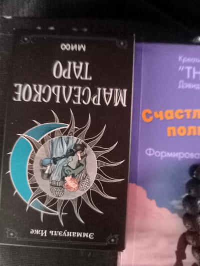 The image shows two books. The book on the left has a black cover with white text. The title appears to be "МАРСЕЛЬСКОЕ ТАРО" which translates to "MARSEILLE TAROT." Below this, it says "ЭЖИ ЧЕЛНЕШШЕ" which is likely the author's name. The cover also features a circular illustration with a crescent moon and a sun-like motif, containing a drawing of two figures.

The book on the right has a purple cover with orange and white text. The orange text reads "Счасть" which translates to "Happiness." Below that, there is orange text that reads "поли" which is likely part of a larger word or phrase. White text above the orange text includes "TH" and what appears to be "Креоты" and "Дэвид".

The books are positioned on a dark surface, possibly fabric. There is no indication of the city of Kashino, Russia, or any specific urban environment in the image. The image is a close-up shot focusing on the book covers.