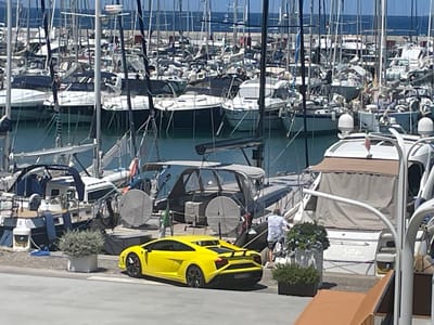 The image depicts a sunny day at a marina in Punta Ala, Italy, with numerous sailboats and motorboats docked in clear blue water. In the foreground, a bright yellow sports car is parked on a paved area adjacent to the docks. Behind the car, a person in a patterned shirt and shorts is standing near a planter box filled with greenery. Several boats are visible, including a covered motorboat with a tan canopy and a white hull, and other vessels with various sails and structures. The background is filled with the masts and rigging of many sailboats. The overall scene conveys a sense of luxury and leisure associated with a coastal resort.