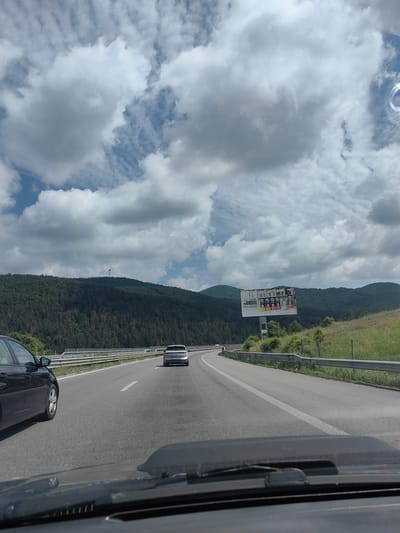 The image depicts a view from a vehicle driving on a highway. The highway curves to the left, flanked by a metal guardrail on the right and a line of trees beyond it. A dark-colored car is visible in the left lane, and a white SUV is ahead in the right lane. In the distance, rolling hills covered in dense green forest rise, with a tall, thin antenna-like structure visible on the crest of one of the hills. A billboard advertising Bialetti coffee products stands on the right side of the highway, against the backdrop of the hills. The sky is filled with a dramatic mix of dark, heavy clouds and patches of bright blue sky, suggesting a day with changing weather conditions. The scene appears to be located near Potop, Bulgaria, given the mountainous terrain and vegetation commonly found in the region.