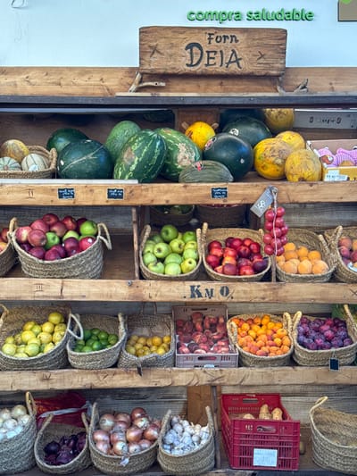 The image depicts a well-stocked produce display at a market stall or shop in Deià, Spain. The display is constructed from multiple horizontal wooden shelves supported by a wooden frame against a light-colored, likely white, wall.

**Subjects/Objects:**
*   **Top Shelf:** Features large gourds and melons, including watermelons and cantaloupes, primarily in green and yellow hues.
*   **Upper Middle Shelf (First Row of Baskets):** Contains numerous wicker baskets filled with various fruits. From left to right: red and green apples in a large basket, green apples in a medium basket, peaches/nectarines in a medium basket, red grapes hanging from a hook, apricots/oranges in a medium basket, and plums/dark fruits in a medium basket. Small black price tags are affixed to the wooden shelf below these baskets, with some visible text such as "3,95 €". A wooden sign above this shelf reads "ForN DEIA" in carved letters, with a small star symbol. Above this, on the wall, green raised letters spell "compra saludable".
*   **Lower Middle Shelf (Second Row of Baskets):** Holds more wicker baskets. From left to right: yellow lemons/citrus, green limes, more yellow lemons/citrus. A "KM 0" stencil is visible on the wooden support beam below this row. A grey plastic crate containing peaches is also present.
*   **Bottom Shelf:** Contains wicker baskets filled with onions (red and yellow varieties) and garlic bulbs. To the right, a red plastic crate holds potatoes.

**Environment:** The setting appears to be an outdoor or semi-outdoor market stall due to the natural light and rustic wooden construction. No people are visible within the frame.