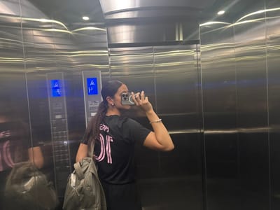 The image displays the interior of a brushed stainless steel elevator located in Santa Catarina, Mexico. The reflective walls feature horizontal light streaks from overhead illumination. On the left wall, two elevator control panels are visible, both displaying a blue "4" with an upward arrow; one panel also indicates "P". A female individual is observed in reflection, positioned to take a selfie with a smartphone held in her right hand and angled towards her reflection. She has dark hair styled in a ponytail. She is wearing a black short-sleeved shirt with pink lettering on the back, distinctly showing "OF" and partially "TEAM" above it. A multi-beaded bracelet is on her right wrist, and a small stud earring is visible in her right ear. A light-colored backpack is slung over her left shoulder. A faint, distorted reflection of the subject, including the backpack and a pink-accented top, is present on the far left.
