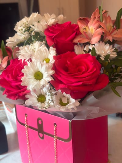 A bouquet of flowers is arranged in a bright pink, purse-shaped box. The bouquet features several large red roses, smaller white daisies with green centers, and some delicate pink and orange lilies. The box has a quilted pattern on its surface and is adorned with gold chains and a gold clasp that has a heart cutout. The arrangement is presented indoors, with a softly lit background that suggests a domestic setting. The specific location in Zonalnaya Stantsiya, Russia is not discernible from the visual information provided in the image.