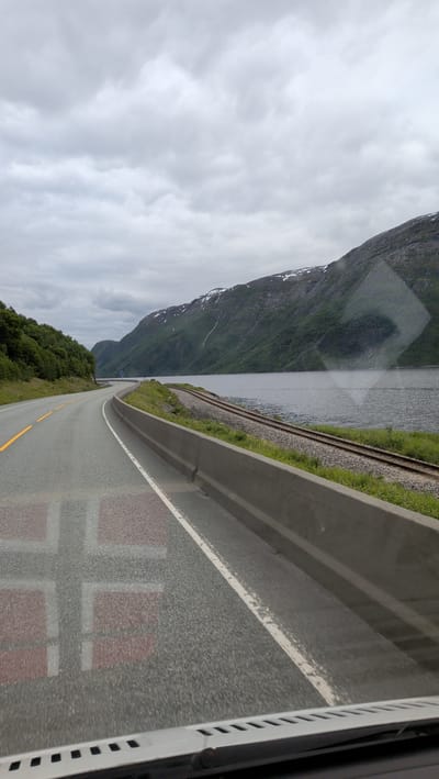 A highway runs alongside a body of water and a railway track. Mountains, partially covered in snow, rise in the distance. A partial reflection of the Norwegian flag is visible on the road. The sky is overcast with heavy clouds.