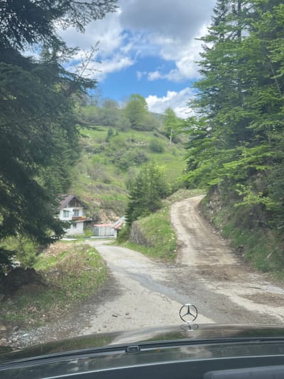 The image, taken from inside a vehicle (likely a Mercedes-Benz, judging by the hood ornament), shows a rural scene near Drianovo, Bulgaria. A narrow, unpaved road winds down a hillside. The hillside is covered in green vegetation, including trees and grass. A small house with a red roof is visible further down the hill. The sky is partly cloudy.