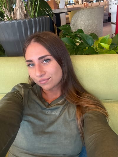 A young woman with long, brown hair and blue eyes is shown in a close-up, vertical, selfie-style shot. She is wearing a long-sleeved, olive-green t-shirt with the word "FREDDY" visible in a vertical line on the left chest area. She is lying down on a lime-green couch. The background includes indoor plants with lush green leaves and a glimpse of what appears to be shelving with decorative items. The lighting suggests it could be daytime. The woman's expression is relaxed and she appears to be smiling slightly. The setting is indoors, possibly a home or a cafe with a natural, relaxed ambiance.