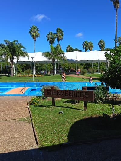 The image depicts a scene at the Parque Termal Daymán in Termas del Daymán, Uruguay. A turquoise-blue swimming pool is in the foreground, with several people inside. A wooden sign with "BIENVENIDOS" (Welcome) in carved letters stands on the grassy bank beside the pool. Behind the pool, a white tensile structure covers a grassy area with palm trees. People can be seen walking and relaxing under the structure. The sky is blue with a few scattered clouds. The ground around the pool is a mixture of concrete walkways and green lawns with some small flowerbeds.